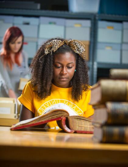 Female student looking at old hardback book with another female student in the background