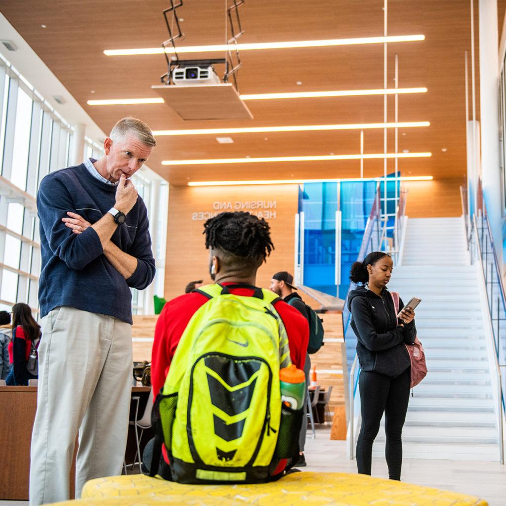 A professor talking to a student in the common area of the nursing and health sciences building.