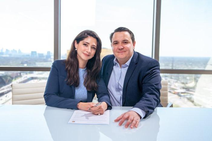 Couple poses for photo in front of glass walls with Dallas cityscape outside. Table in front of them includes their signed gift agreement.