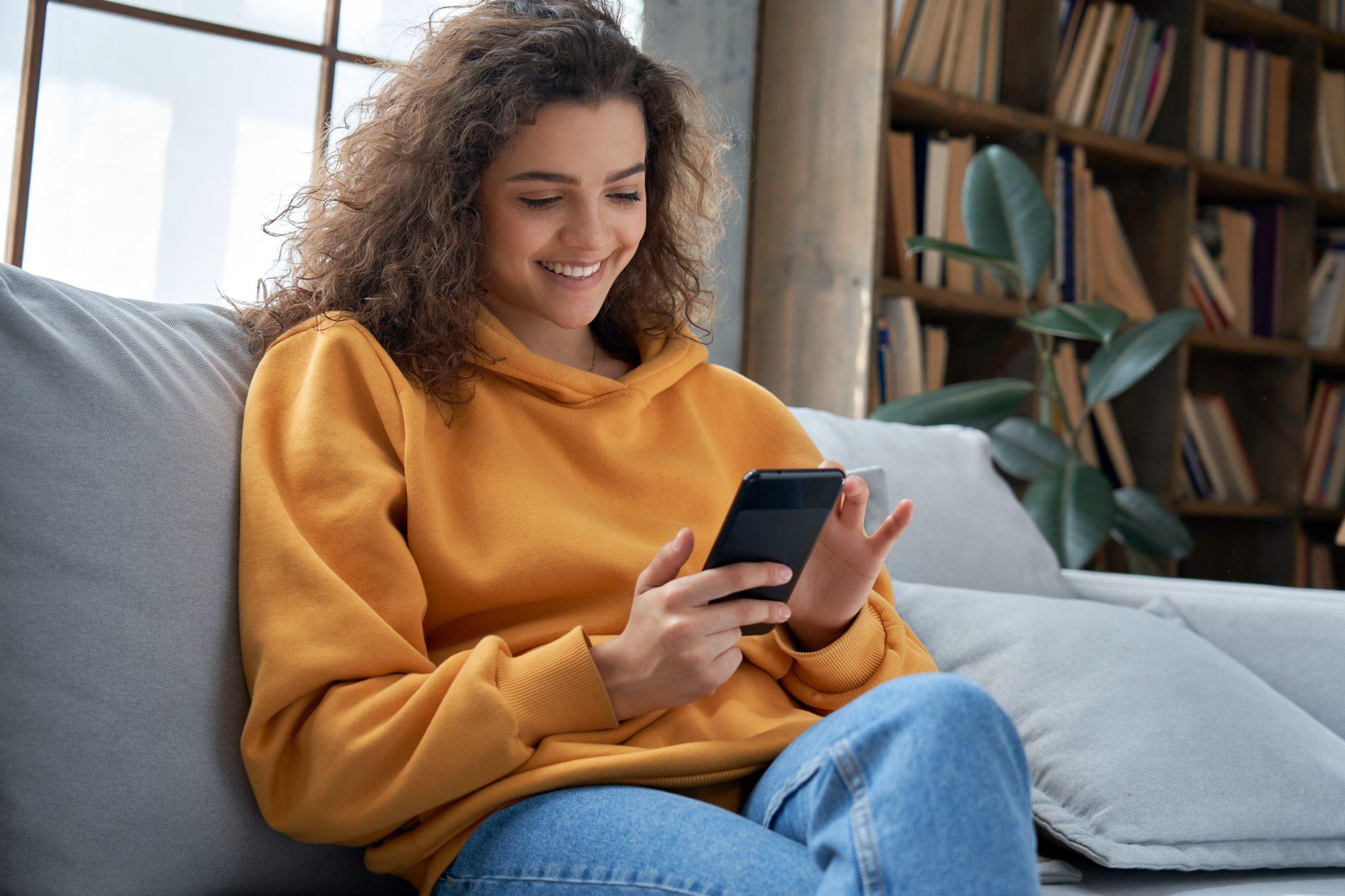 A female on the couch looking through her phone while smiling.